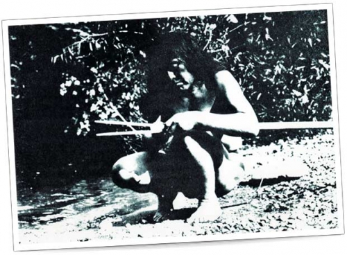 Traditionelle Jagdmethode – Schwarzweiß-Foto Ein Mann sitzt am Ufer eines Flusses und bearbeitet einen Speer. Er ist barfuß und trägt natürliche Kleidung.