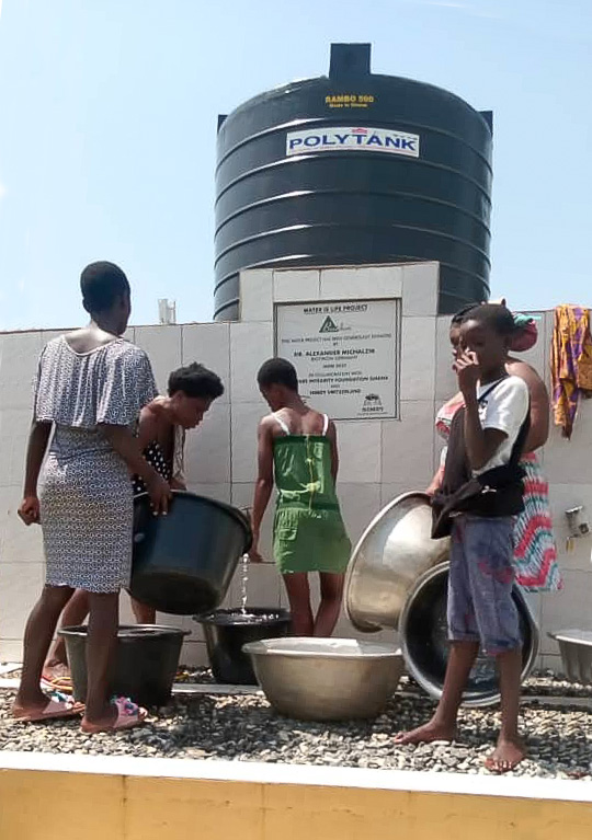 Wassertank – Wasserentnahme in Behältern Gruppe von Kindern entnimmt Wasser aus einem Wassertank, verschiedene Behälter, helle Kleidung, sonnige Umgebung.