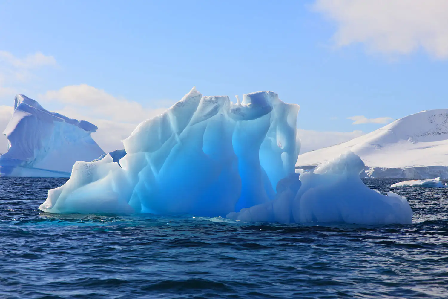 Eisberg Ein blauer Eisberg schwebt im ruhigen Wasser, Umgebung zeigt schneebedeckte Berge und Himmel.
