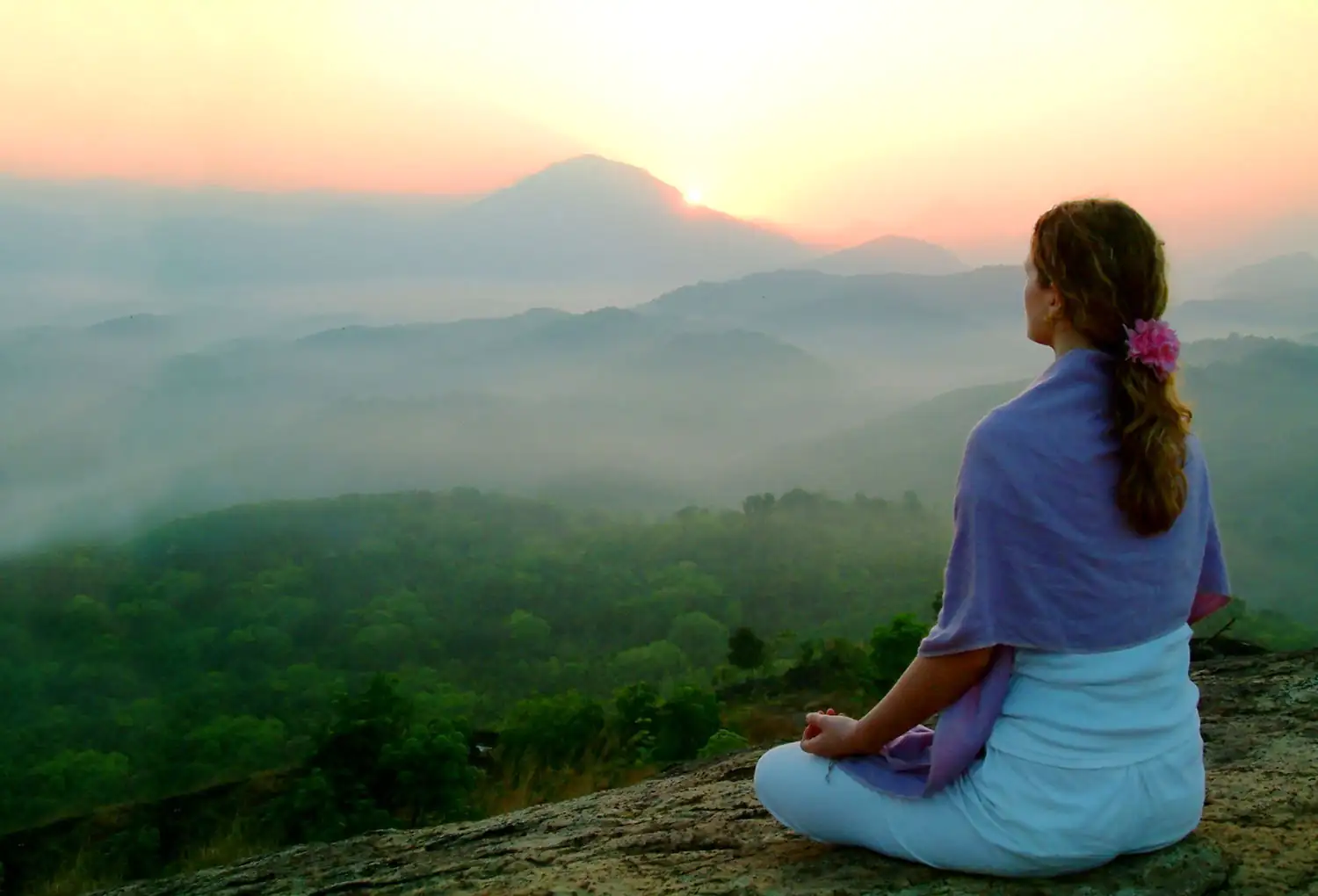 Frau meditiert bei aufgehender Sonne in der Natur Frau sitzt meditierend auf einem Felsen, umgeben von einer nebligen Berglandschaft bei Sonnenaufgang.