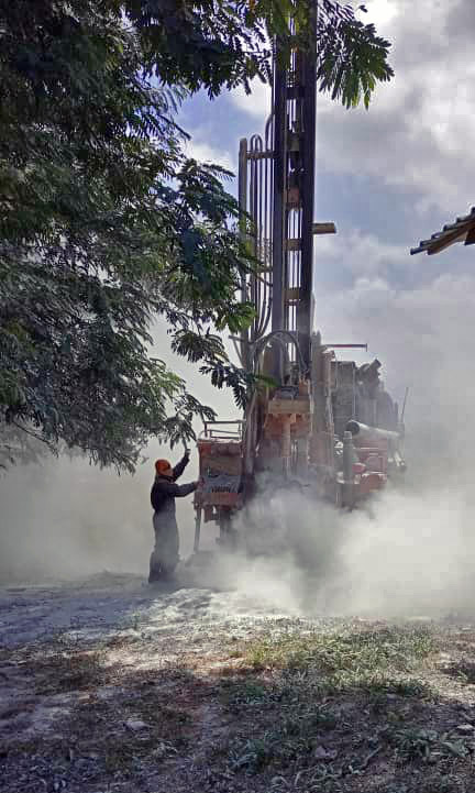 Bohrmaschine – Wasserbohrung vor Ort Arbeiter steuert eine Bohrmaschine in einer staubigen Umgebung, Bäume im Hintergrund.