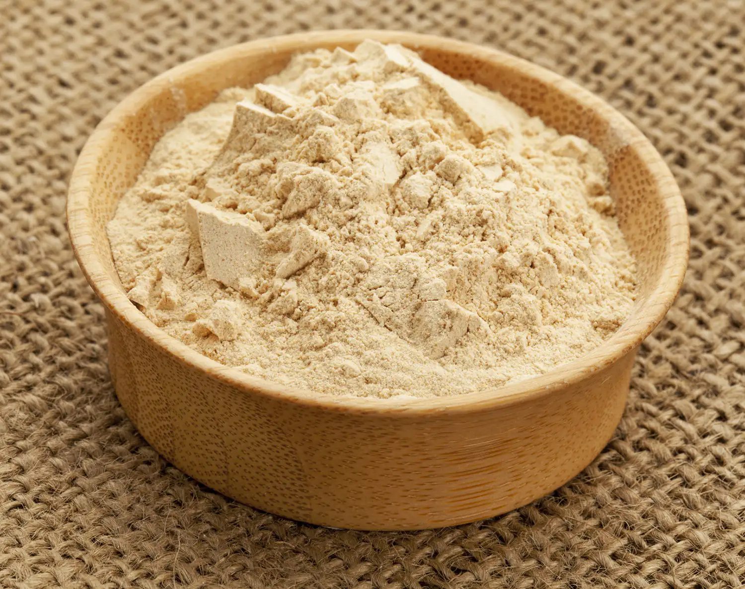 Maca Pulver Fein gemahlenes Maca Pulver in einer Holzschale, beige Farbe, auf einer Jute-Oberfläche.